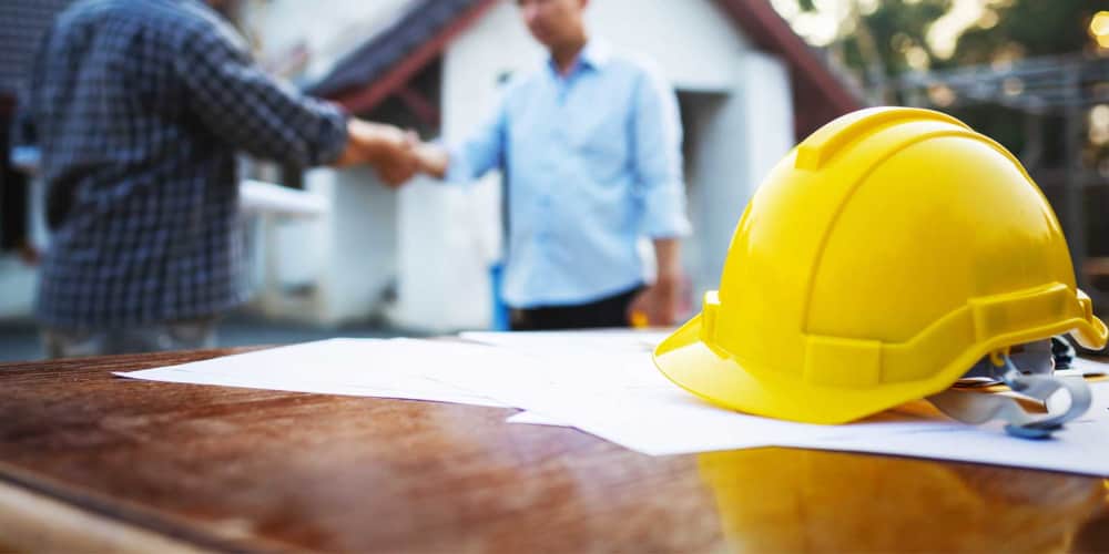 Two workers shaking hands, in the foreground there is a yellow hard hat. 