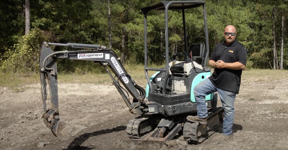 A DOZR equipment expert standing next to a mini excavator explaining how to operate it