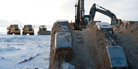 Close up view of metal equipment tracks with two yellow excavators, a bulldozer, and two articulated dump trucks behind it on snow-covered dirt ground.