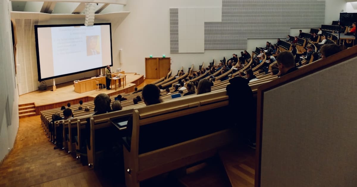 A university lecture hall viewed from the back, students seated in curved rows facing a presentation screen