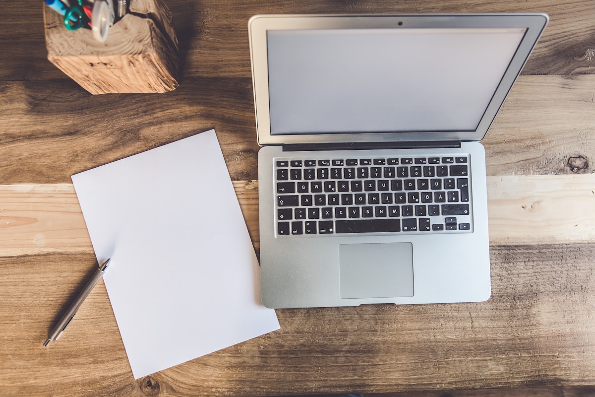 A laptop viewed from above with its blank screen open beside a sheet of blank paper and a pen on a wooden desk