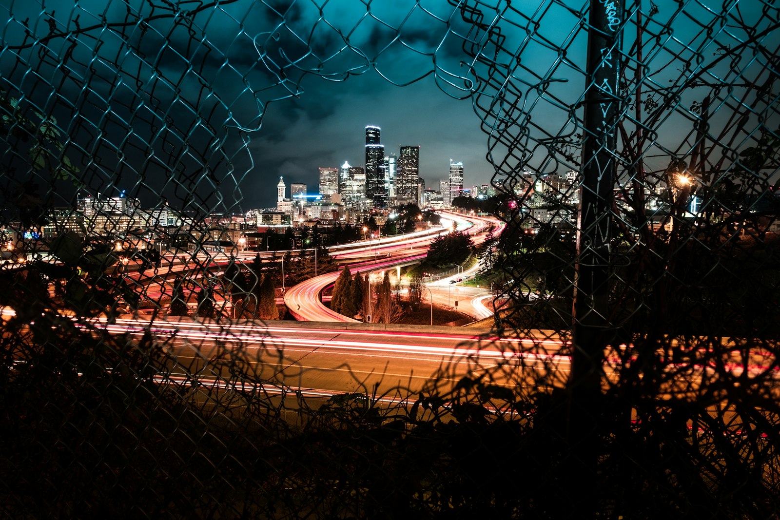 A city skyline and light-trail traffic seen at night through a chain-link fence