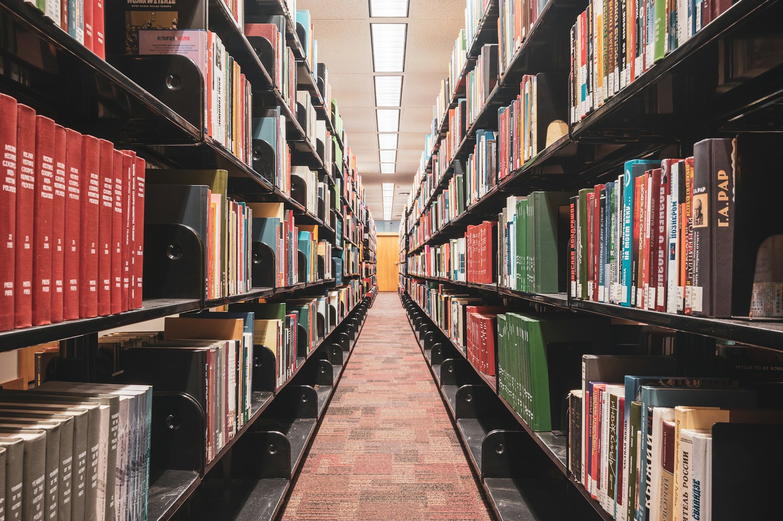 A long aisle between tall library shelves packed with books, spines facing outward under overhead lighting