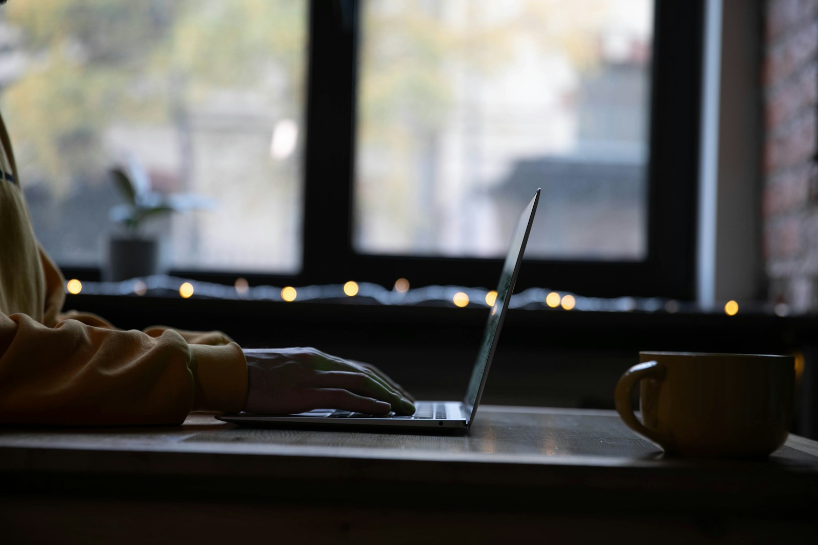 A hand resting on a laptop keyboard at a desk in the evening, with a mug beside it