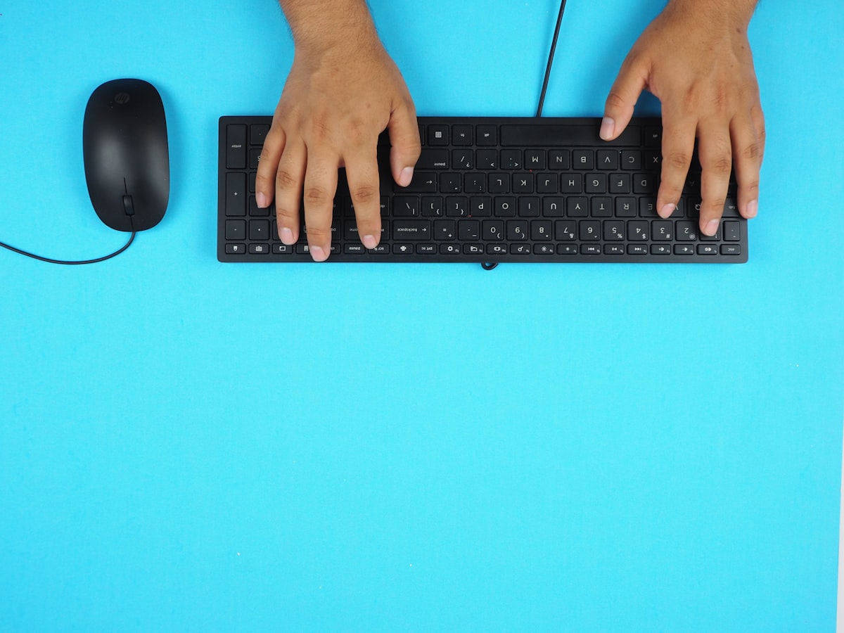 A pair of hands typing on a black keyboard against a plain blue desk surface