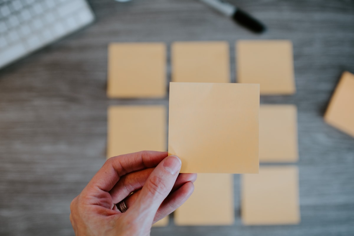 A hand holding a blank sticky note above a grid of other blank sticky notes on a desk
