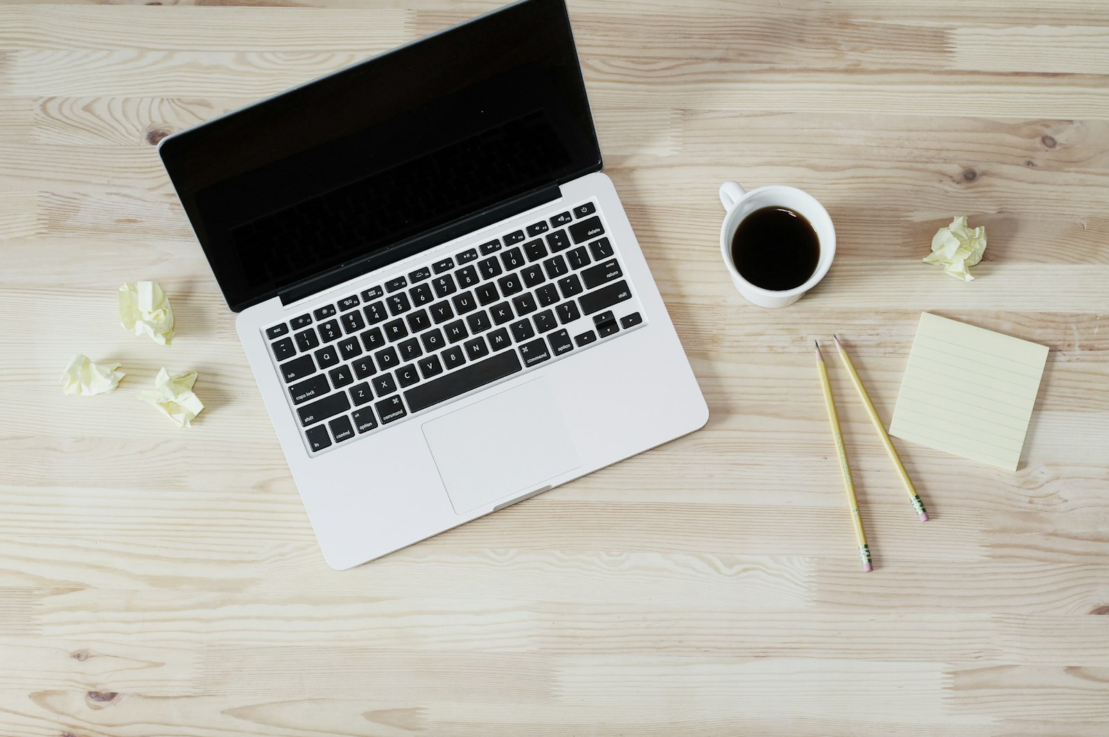 A laptop with its screen off next to a cup of coffee, two pencils, and a blank sticky note pad