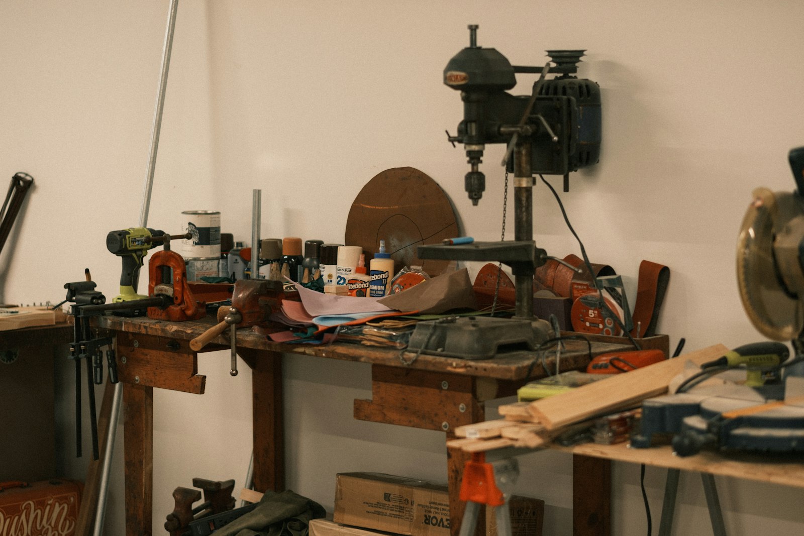 A cluttered workshop bench with hand tools, clamps, and a drill press