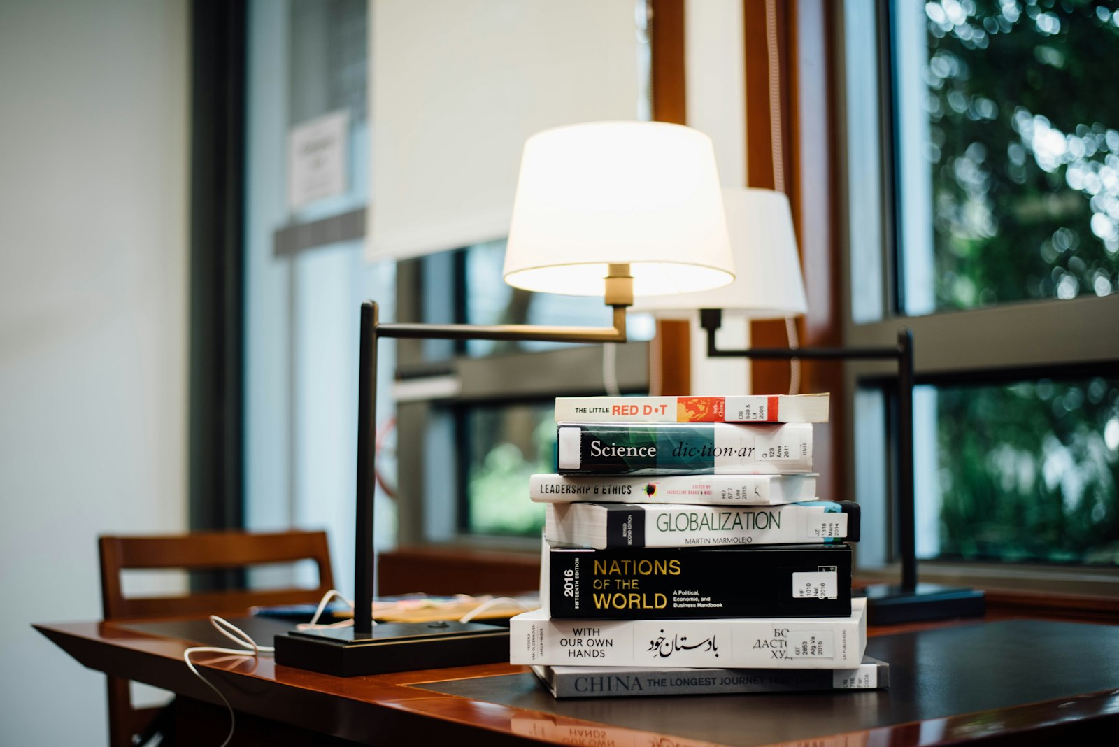 A stack of books beside a lit desk lamp on a wooden desk near a window