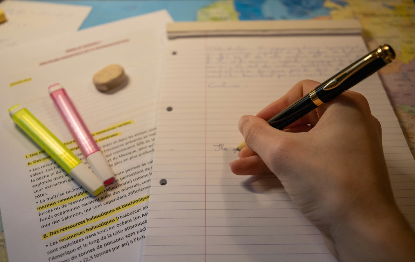 A hand writing notes with a pen beside a printed page marked with yellow and pink highlighter