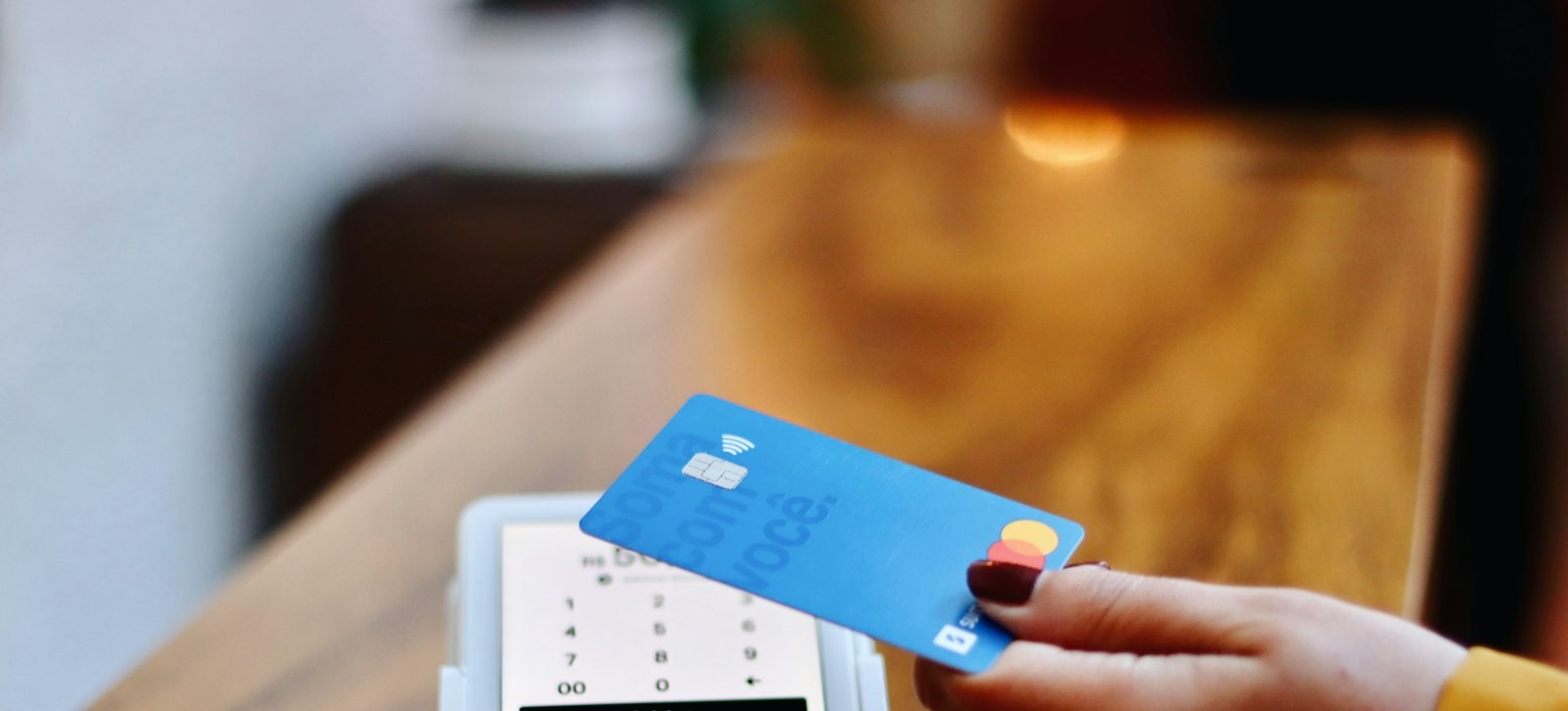 A hand tapping a card against a payment terminal on a shop counter, representing a transaction between a seller and a customer