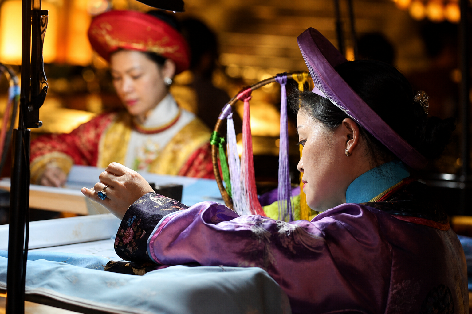 Two women in traditional Vietnamese Ao Dai and hats are engaged in silk weaving, with colorful threads on a loom.