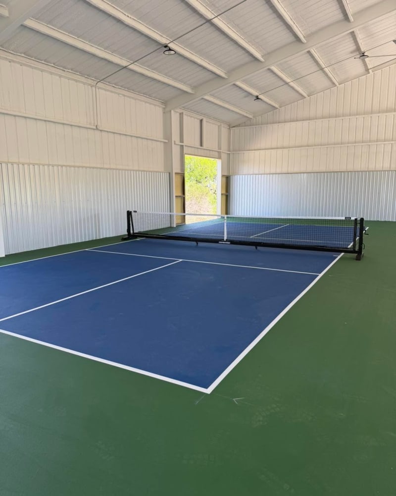 Indoor pickleball court in blue with green surround and a black portable net inside a white metal building, open door to trees
