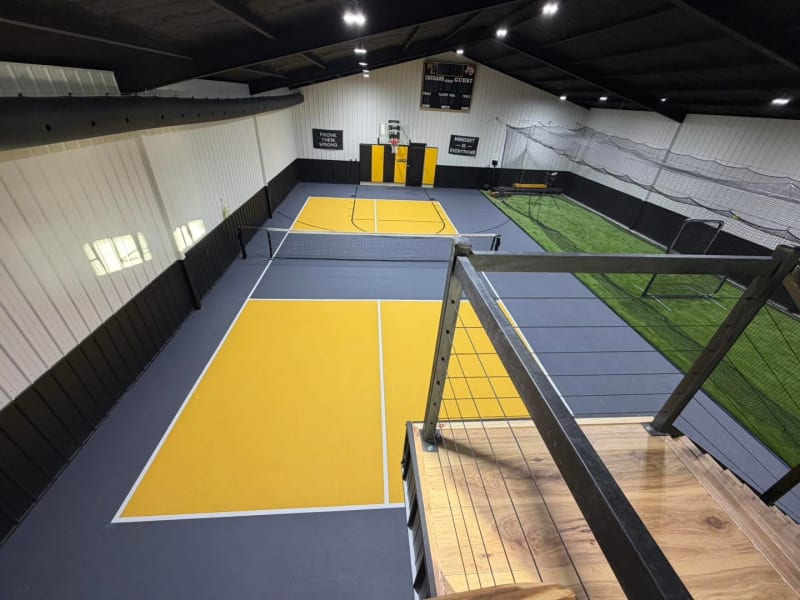 View from a loft rail over an indoor yellow and gray pickleball and basketball court beside a turf batting cage