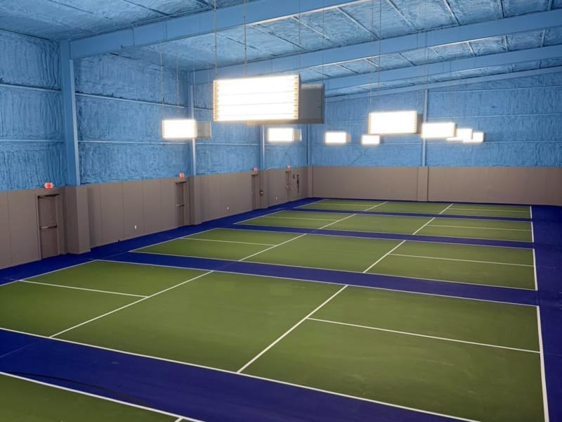 Indoor pickleball facility with a row of green courts and blue borders, white lines, hanging LED lights and blue spray-foam ceiling