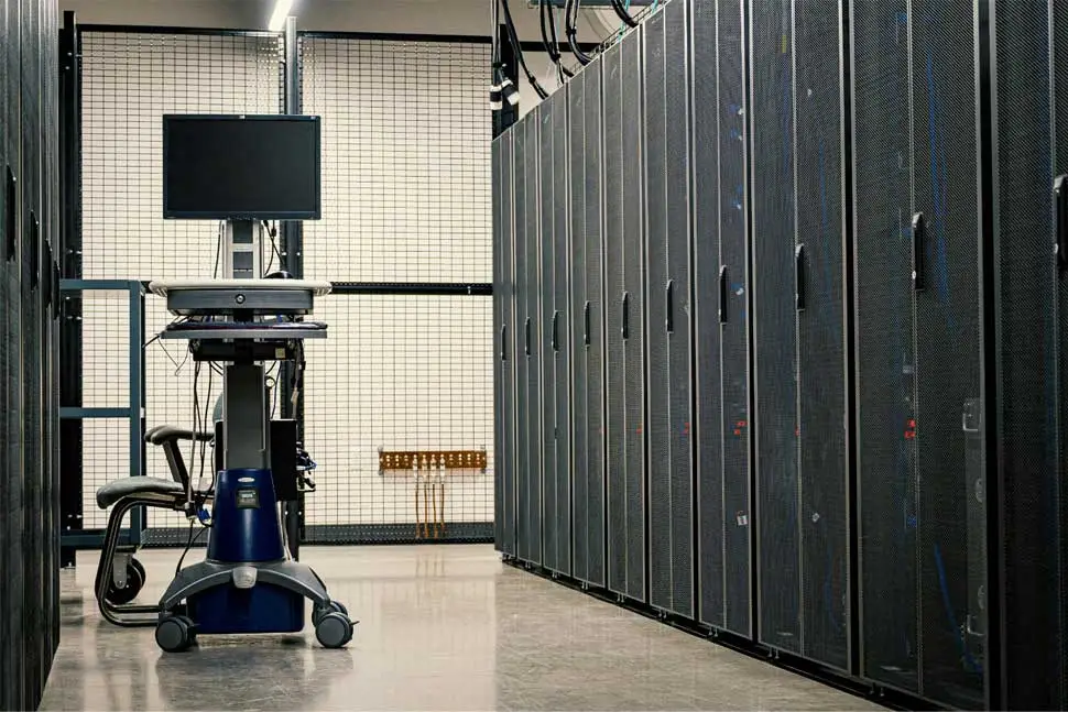 Interior of a modern data center featuring a row of server racks with mesh doors, a movable workstation cart with a monitor and keyboard. The clean and organized setup emphasizes controlled environmental conditions and IT infrastructure management.