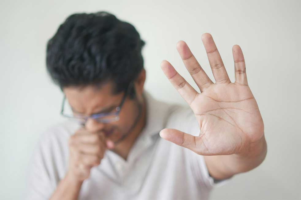 A man wearing glasses coughing or reacting to a bad smell, covering his mouth with one hand while extending the other hand forward in a gesture to stop or avoid something unpleasant.