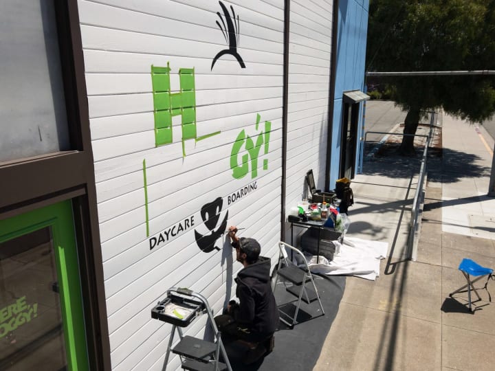 Artistic depiction of a dog daycare facility's logo being meticulously painted on its building's facade, highlighting exceptional mural and sign making talents.