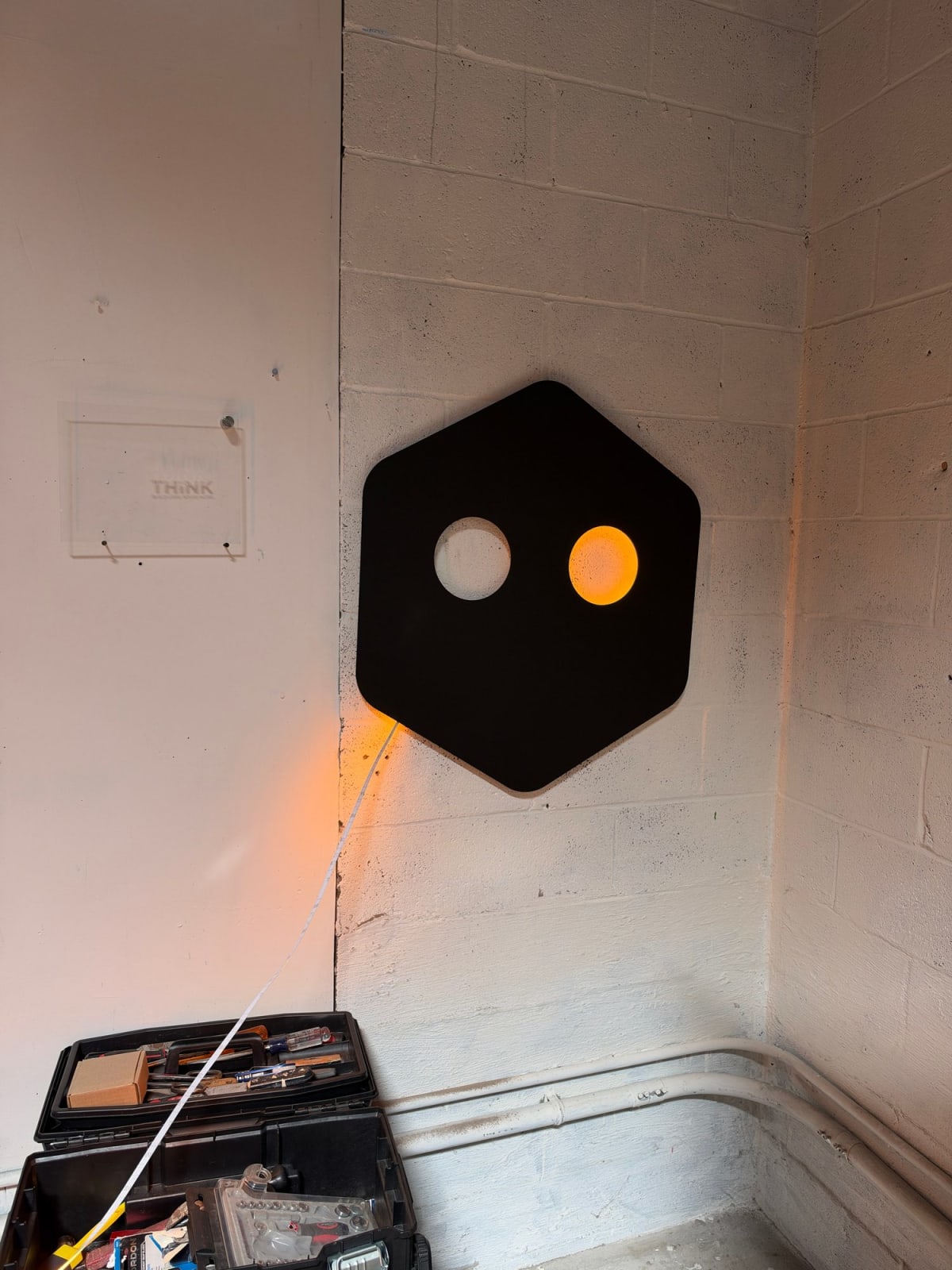 Black custom sign on a workshop wall with its right circular opening glowing amber and test wires visible below