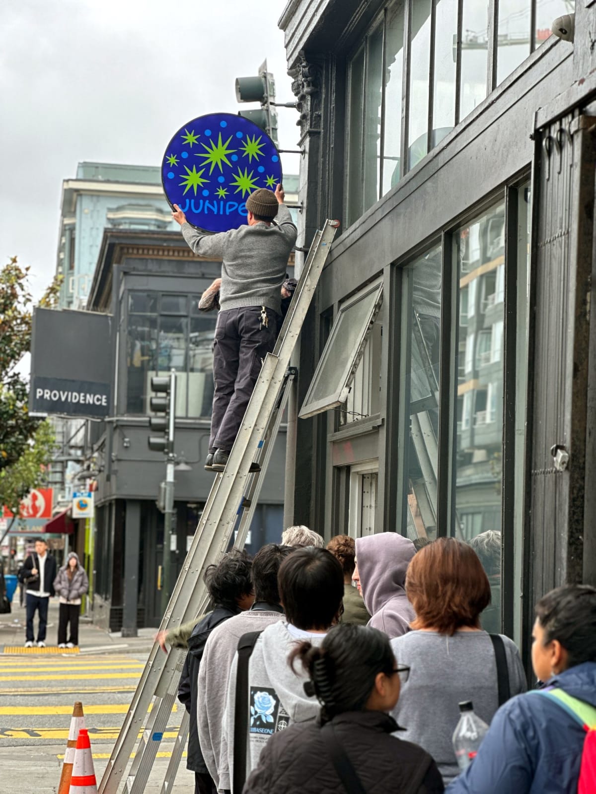 Juniper Cafe blade sign being installed above a sidewalk crowd in San Francisco
