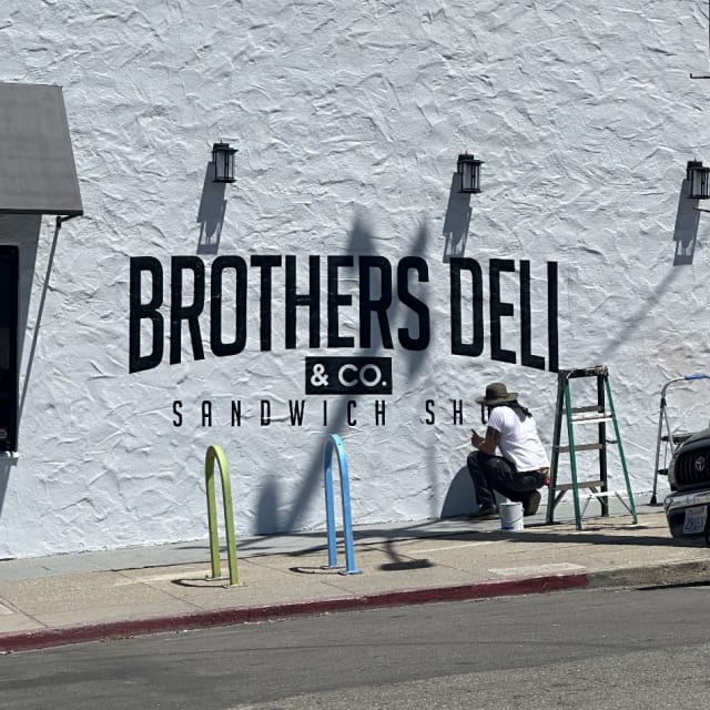 Painter working beneath Brothers Deli lettering on a white exterior wall in Sacramento