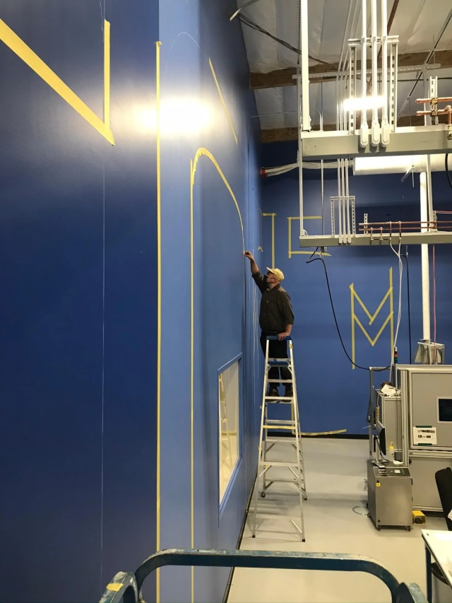 Painter on a ladder following taped letter outlines on a tall blue wall