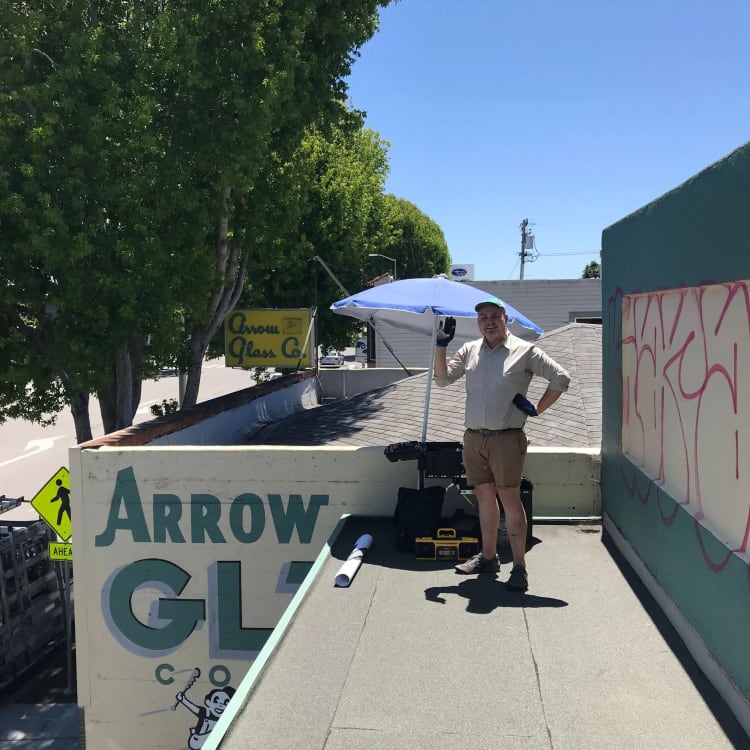 Worker waving from a rooftop beside graffiti and an Arrow Glass Co. sign.
