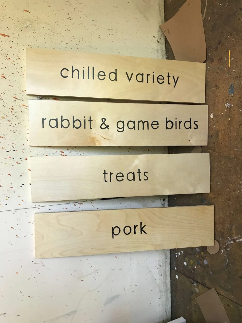 Four unfinished wood category signs labeled “chilled variety,” “rabbit & game birds,” “treats,” and “pork” on a workshop floor.