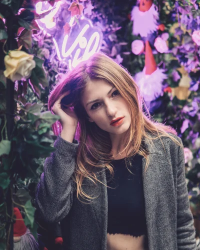 Image shows Maryna Sedin posing thoughtfully amidst a lush backdrop of green foliage and vibrant purple flowers. She's illuminated by a soft pink neon glow that highlights her features. She's wearing a textured grey coat over a black top and navy blue trousers. Her makeup is natural with a striking red lipstick, and she's gently touching her hair, giving the camera a pensive, slightly parted-lip gaze.