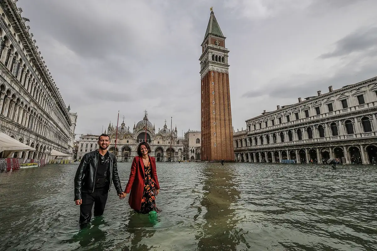 Couple walking through floodwaters in Piazza San Marco, Venice, with St. Mark’s Basilica and the Campanile tower in the background during high tide.