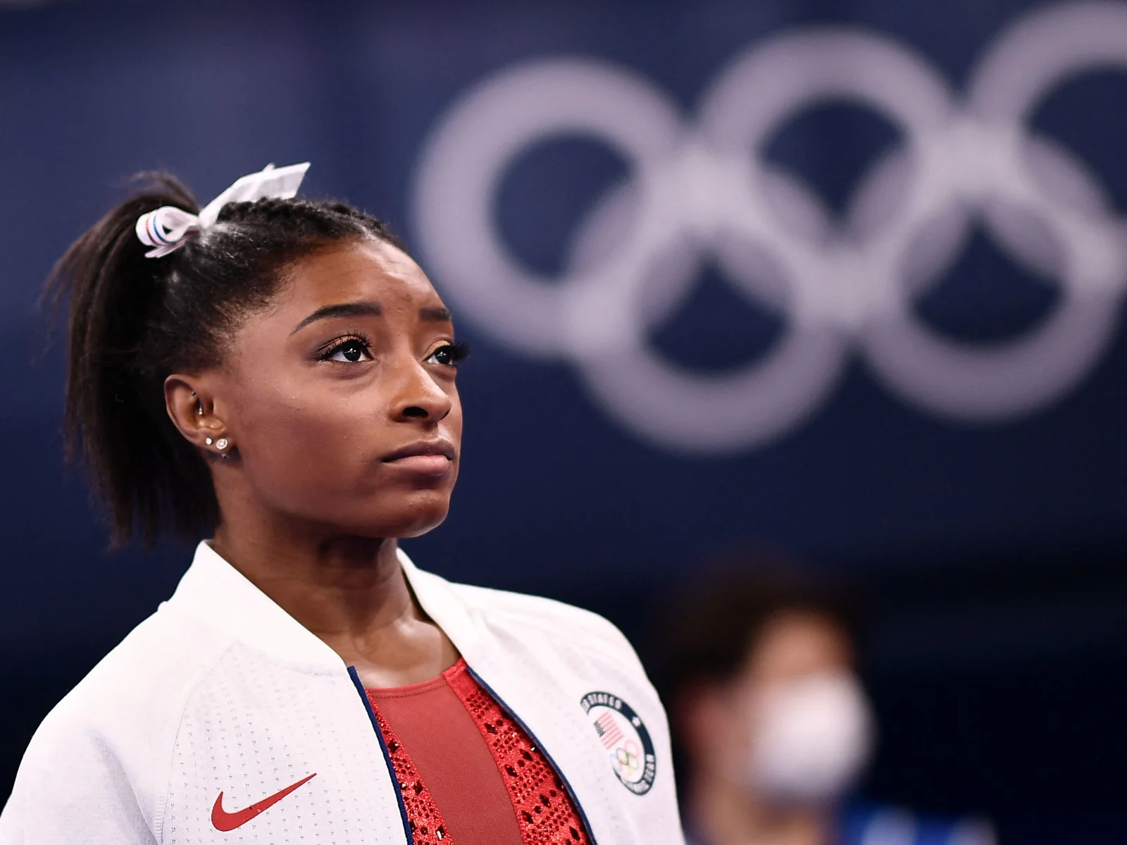 Close-up of a U.S. gymnast wearing a white Team USA jacket with Olympic rings in the background during competition.