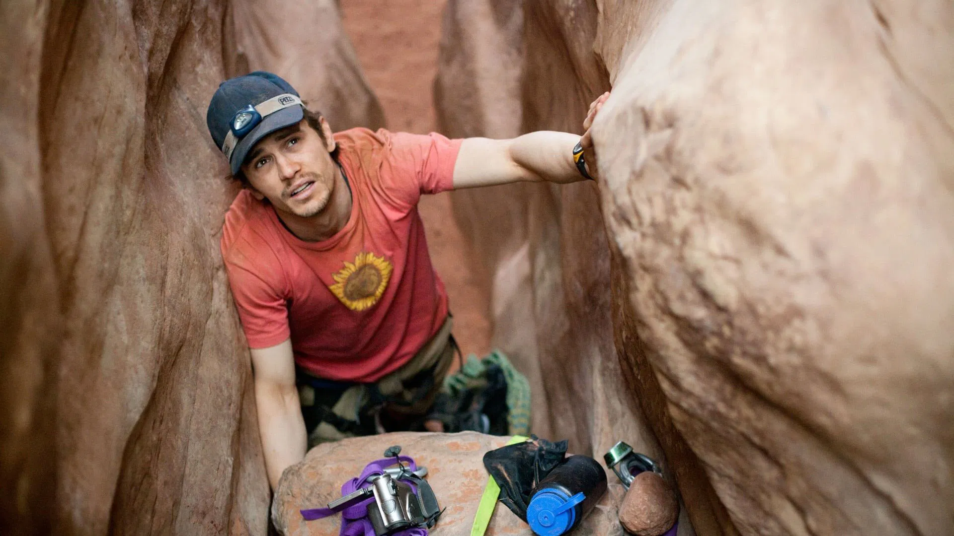 Man trapped between canyon walls with arm pinned under a boulder, wearing red shirt and headlamp, survival scene from 127 Hours.