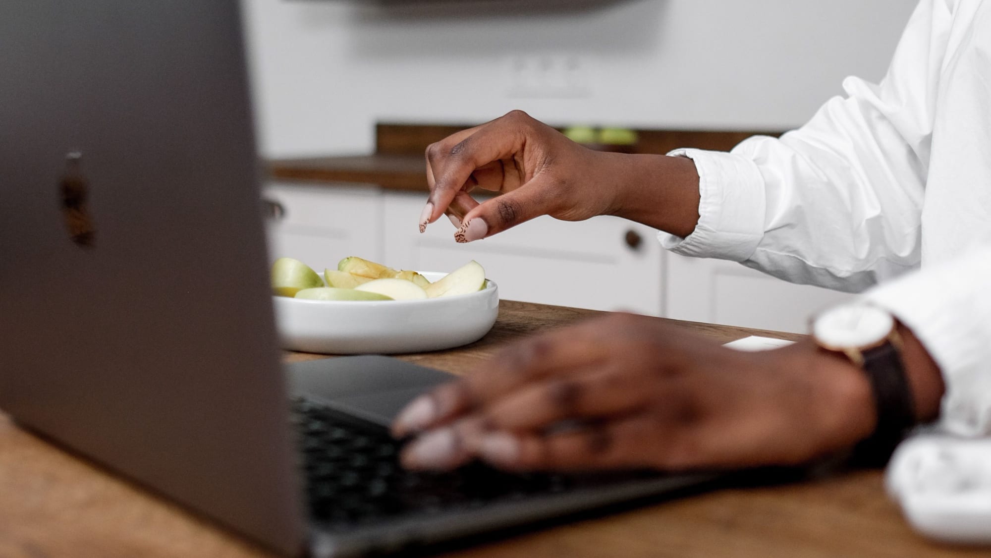 An individual multitasking with a laptop and food on a wooden table indoors.