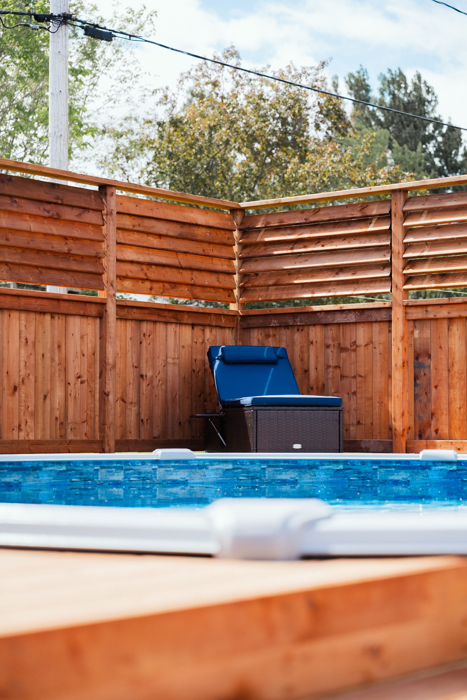 Detail of cedar-toned pool deck boards meeting the pool edge with a poolside lounger and privacy louvers behind, Riverview NB