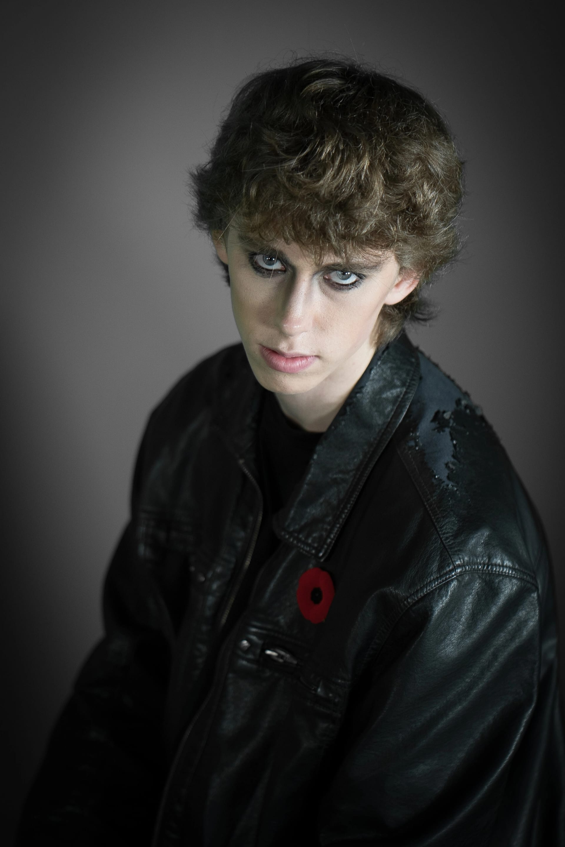 This is a portrait of a person, wearing a black leather jacket with a poppy on it and dark makeup, set against a gray background. The subject is looking directly at the viewer.
