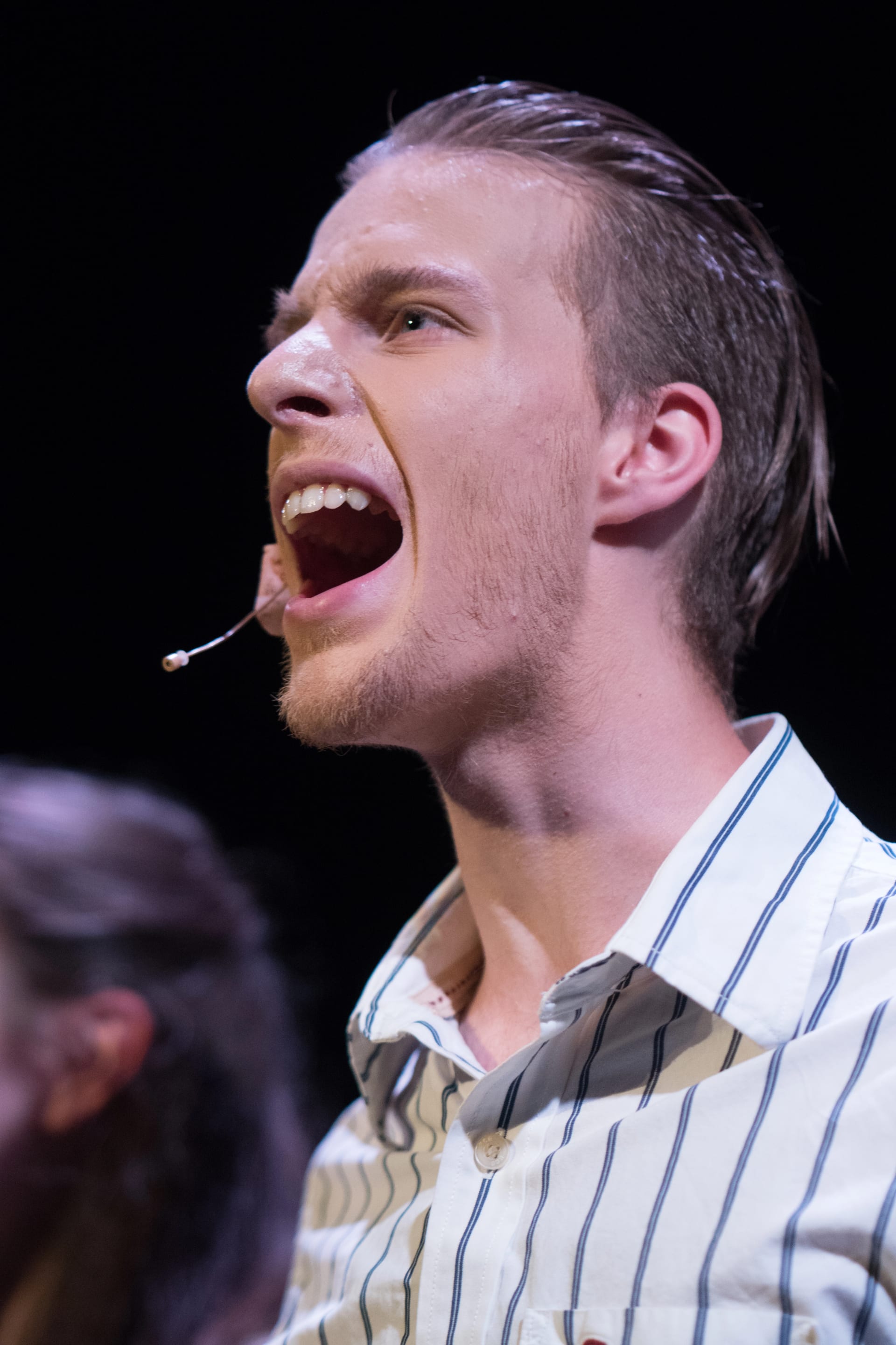 A close-up shot captures a man mid-speech, mouth agape with a look of intensity. The subject is well-lit against a dark background.