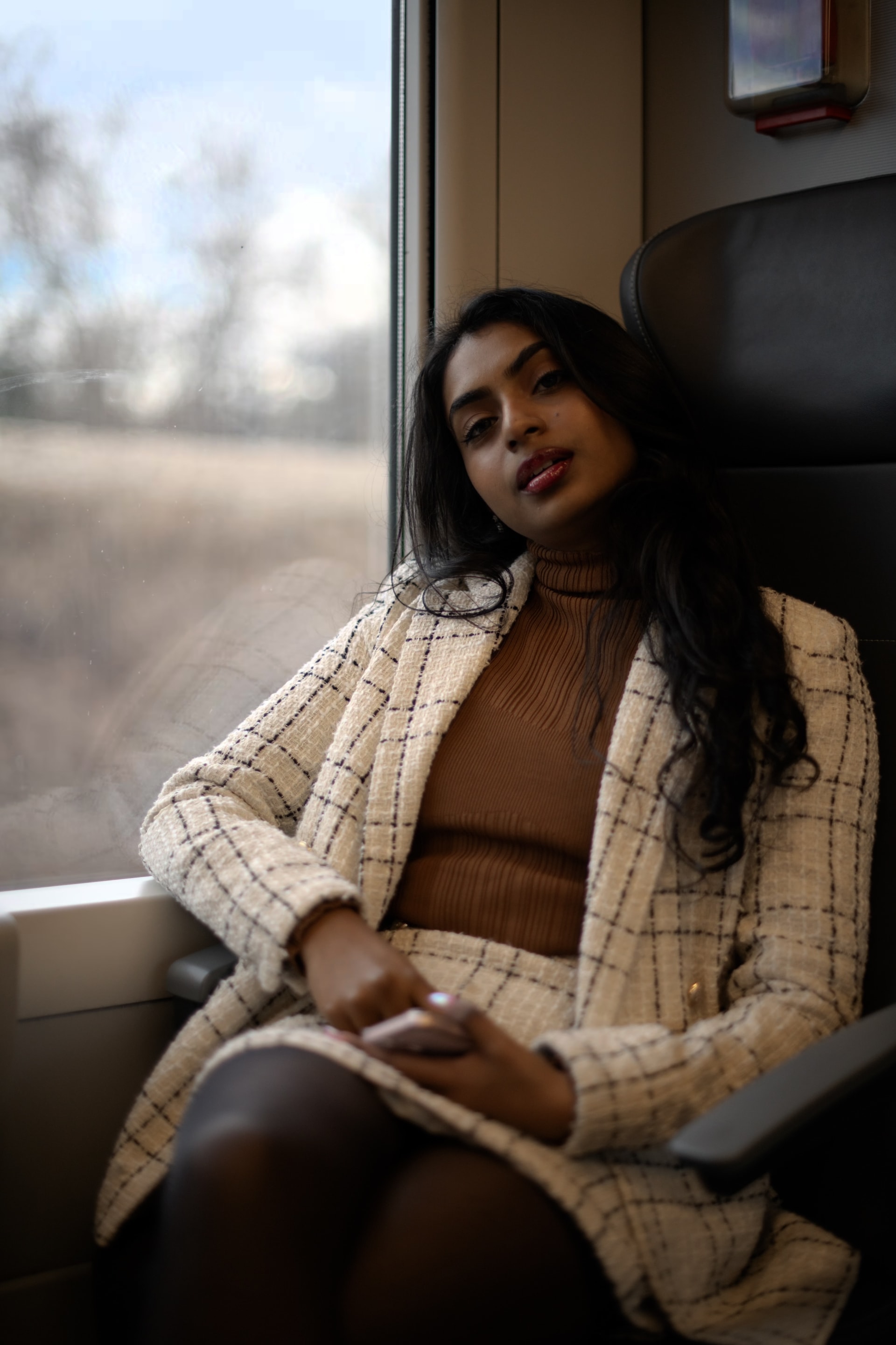 A woman in a blazer and skirt sits on a train, looking at the viewer with a slight smile.