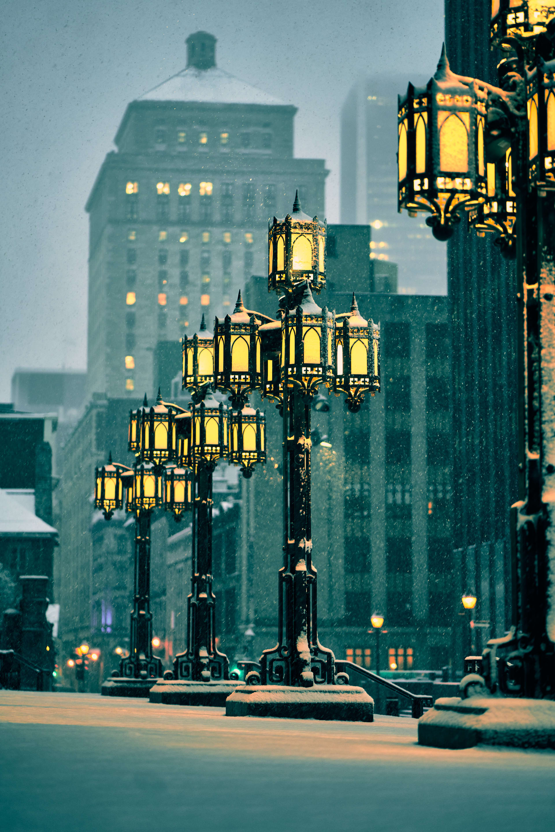 A nighttime winter scene with ornate streetlights illuminated against a backdrop of buildings. Snow falls gently, creating a serene and atmospheric ambiance.