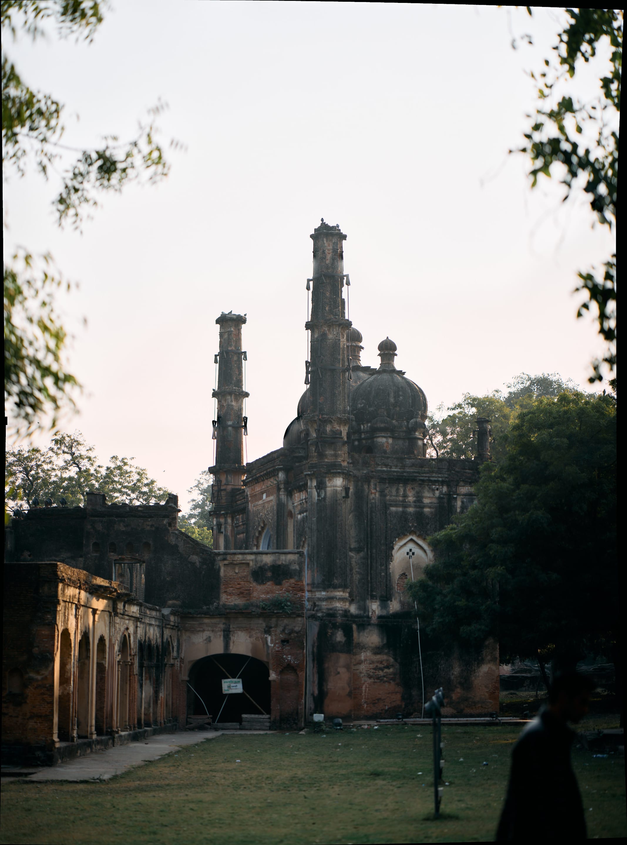 An old building with multiple towers is framed by trees, with a person in the bottom right. The building has a dark color, and some structures like arches can be seen on the sides.