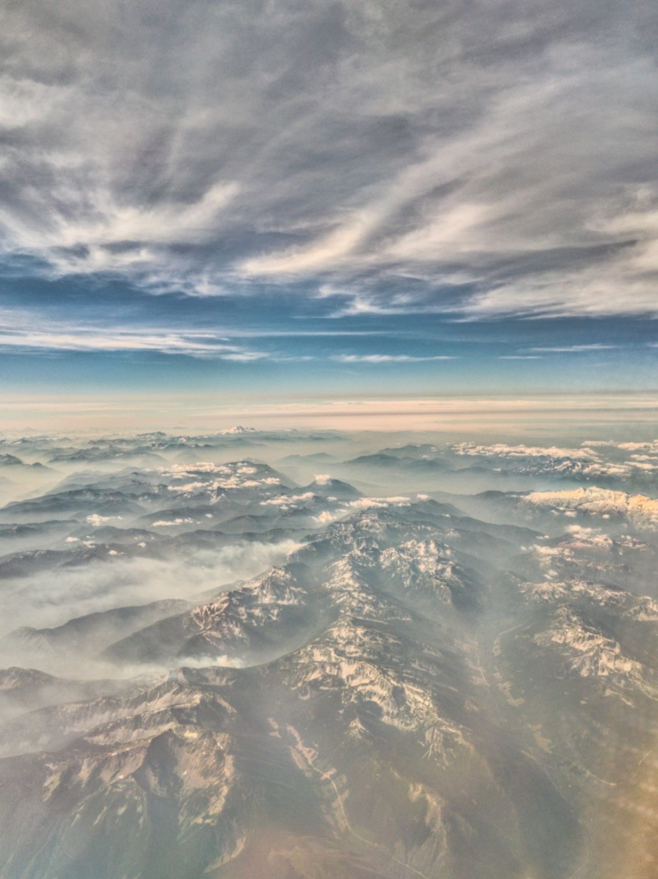An aerial view of a mountain range covered in snow and mist, under a dramatic cloudy sky. The scene captures the vastness and serenity of nature.