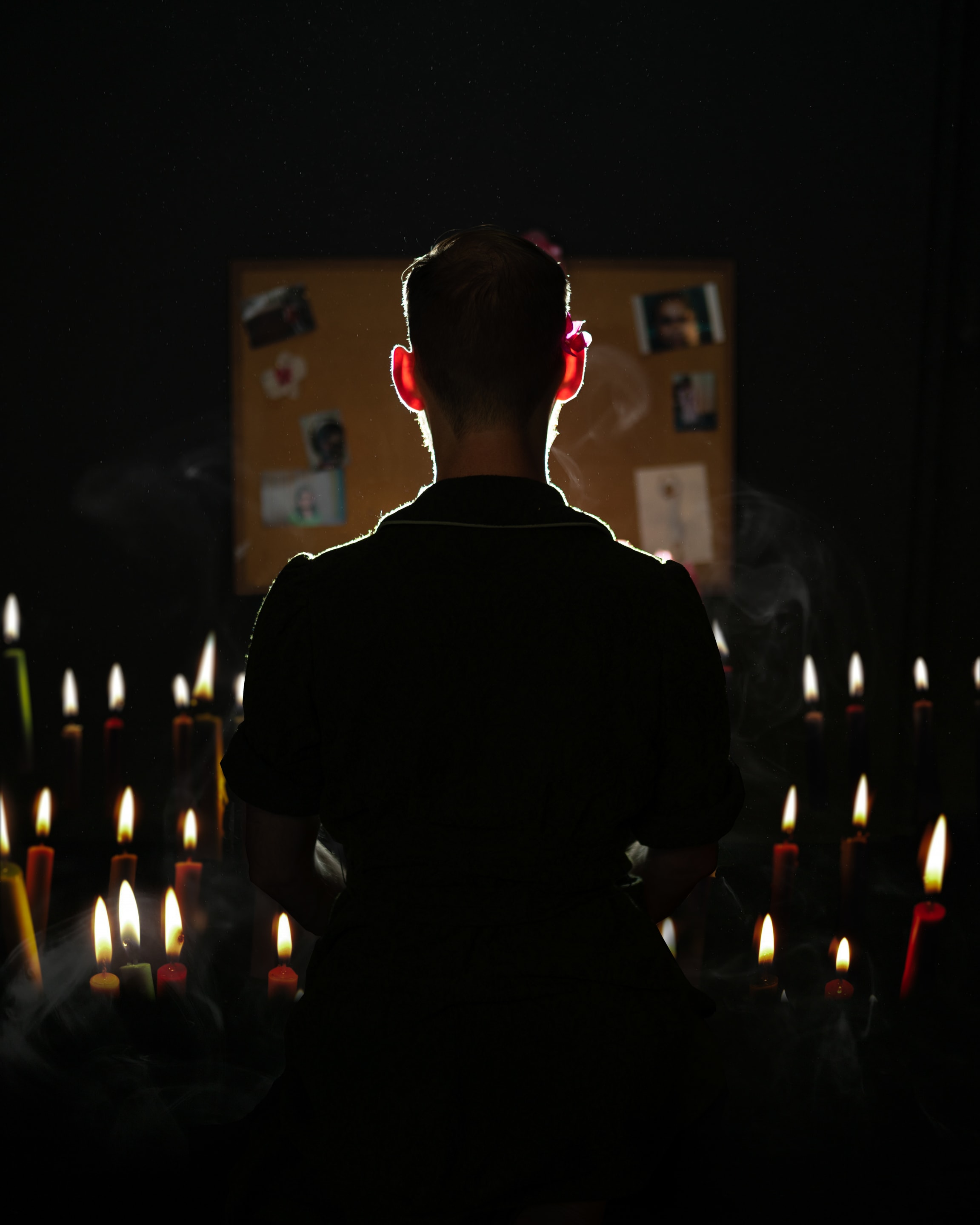 A figure stands before a display of photos and candles. The scene evokes a sense of loss, reflection, and remembrance.