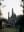 An old building with multiple towers is framed by trees, with a person in the bottom right. The building has a dark color, and some structures like arches can be seen on the sides. blurred out low res