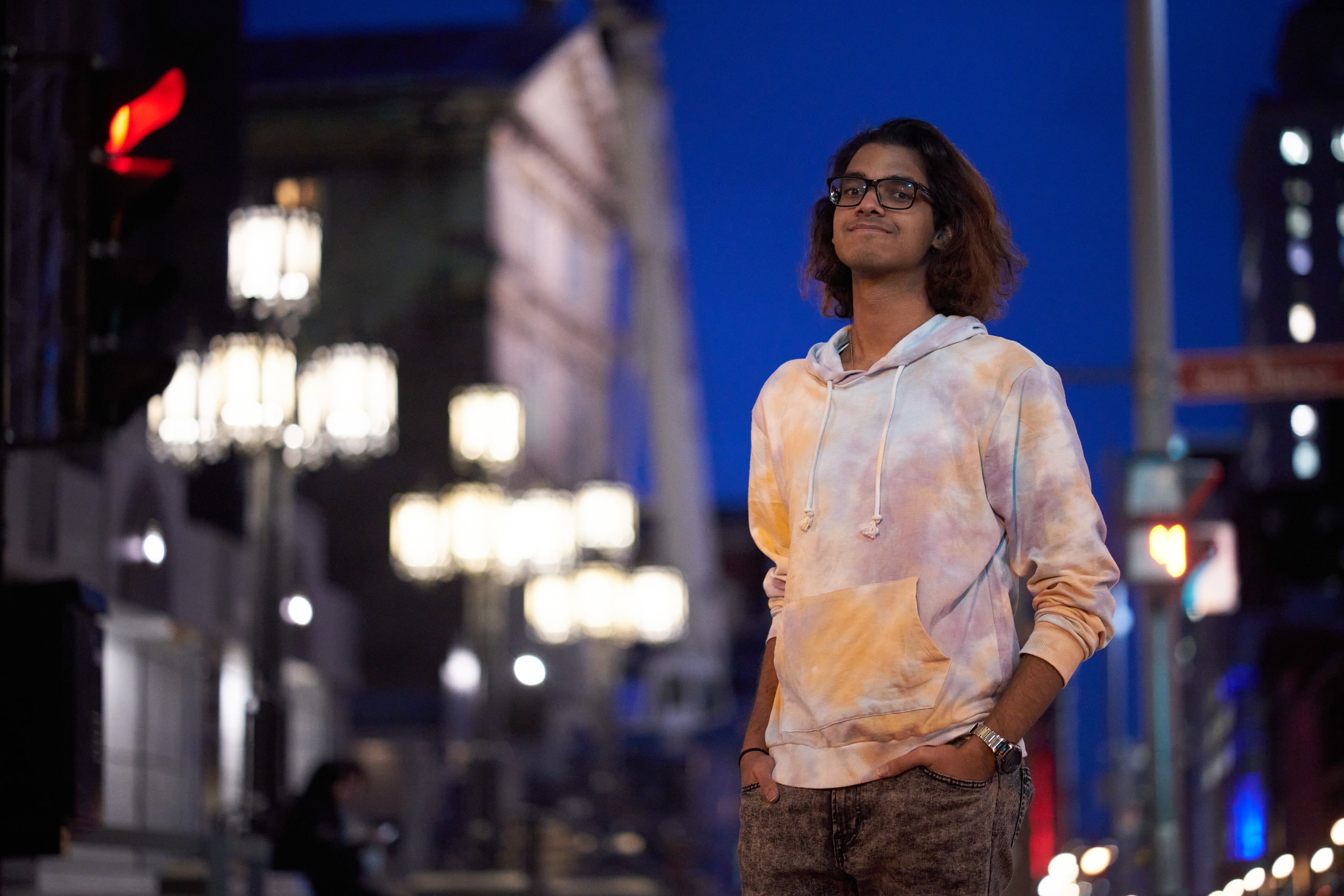 A young man in a colorful hoodie stands confidently on a city street at dusk, surrounded by blurred lights.