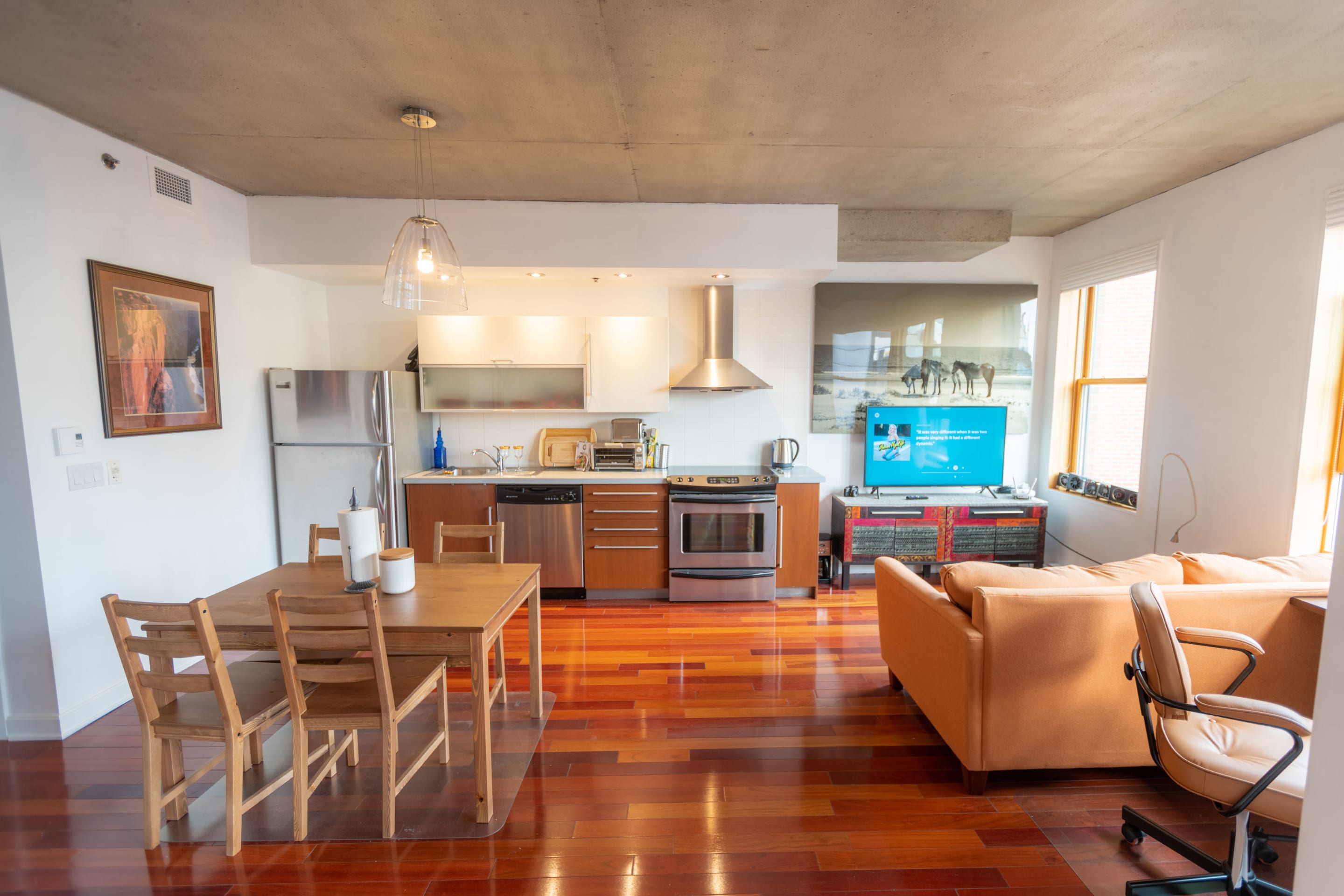 An interior shot of a modern living space, featuring a kitchen, dining area, and living room. The scene is illuminated by natural light.
