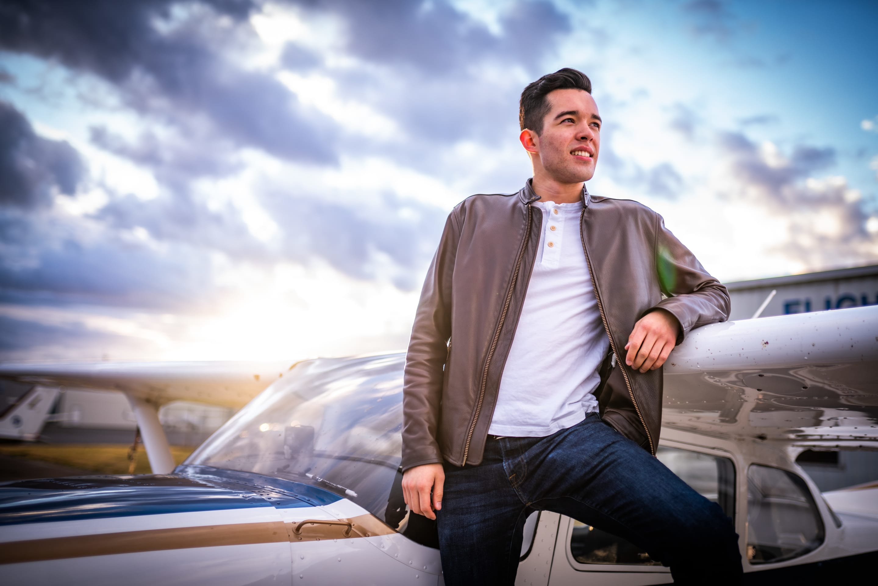 A man leans against an airplane wing, gazing off into the distance. The sky is filled with beautiful clouds and sunset.