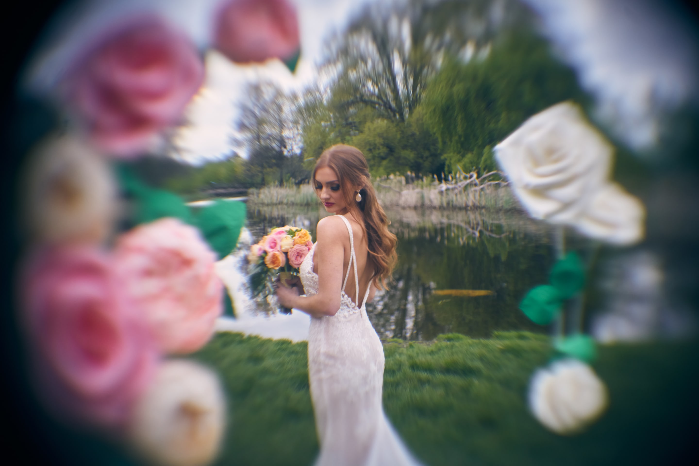 A bride poses by a tranquil lake, surrounded by soft-focus floral elements. The image evokes a romantic and dreamy atmosphere, ideal for capturing the essence of a wedding.