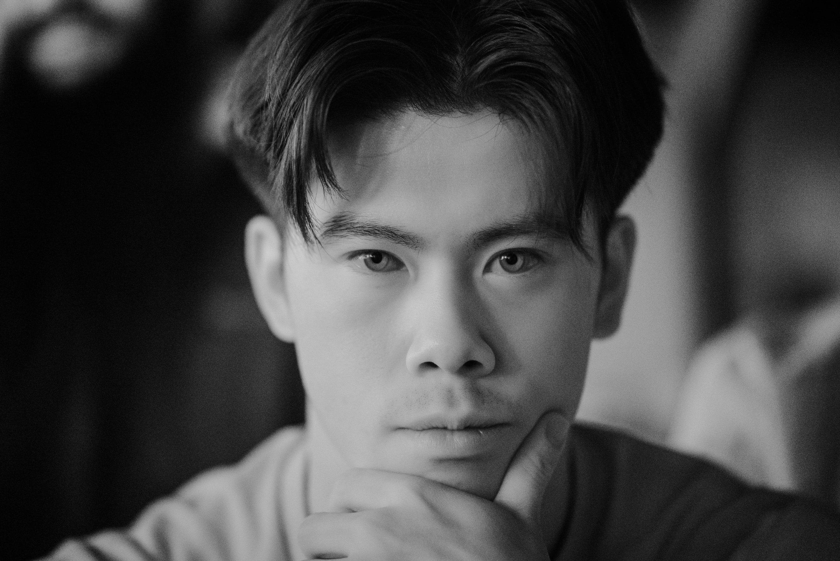 A black and white close-up portrait of a young Asian man. The image focuses on his face, capturing his contemplative gaze.