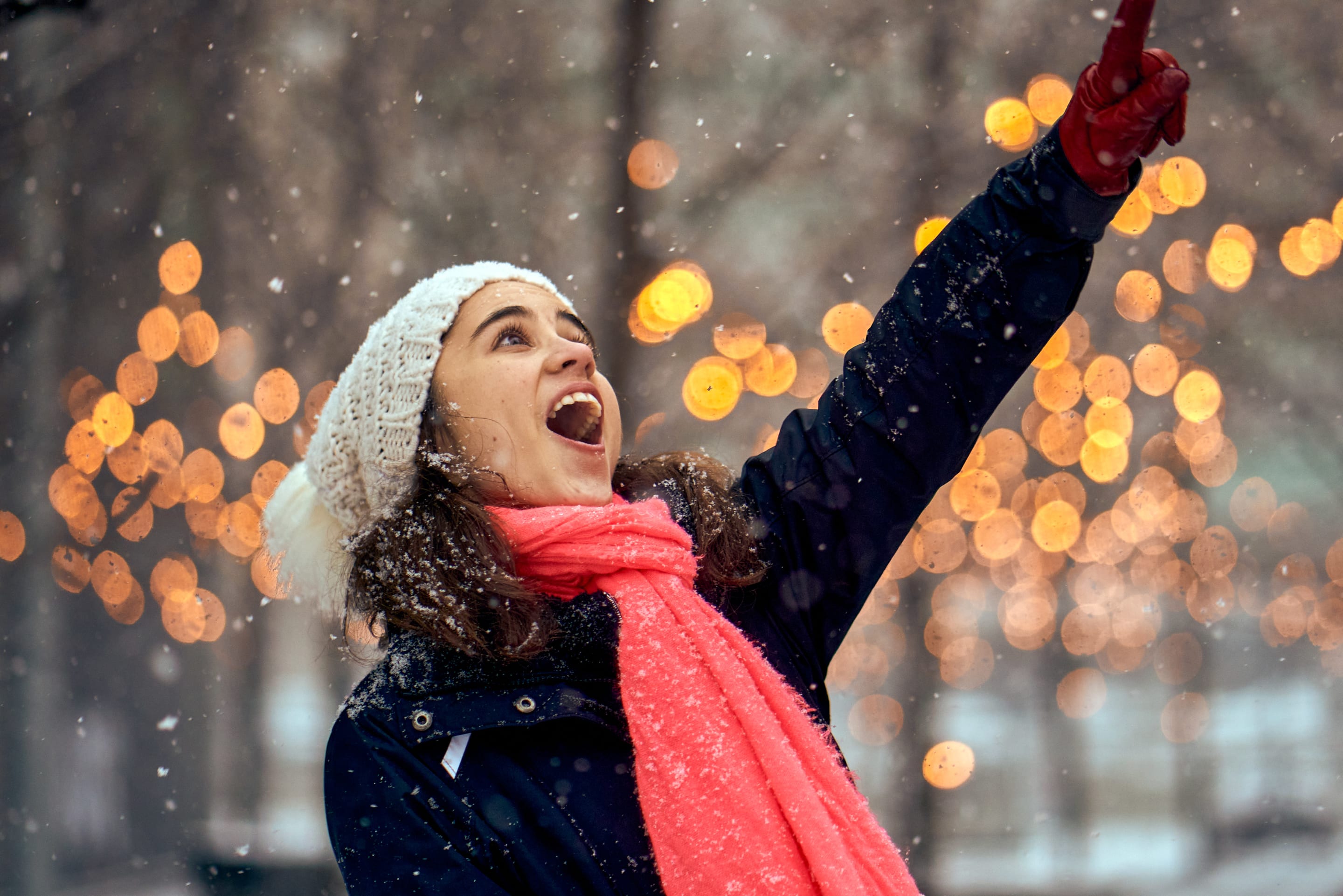 A woman joyfully points upwards while it snows. The background features bokeh lights, enhancing the festive atmosphere.
