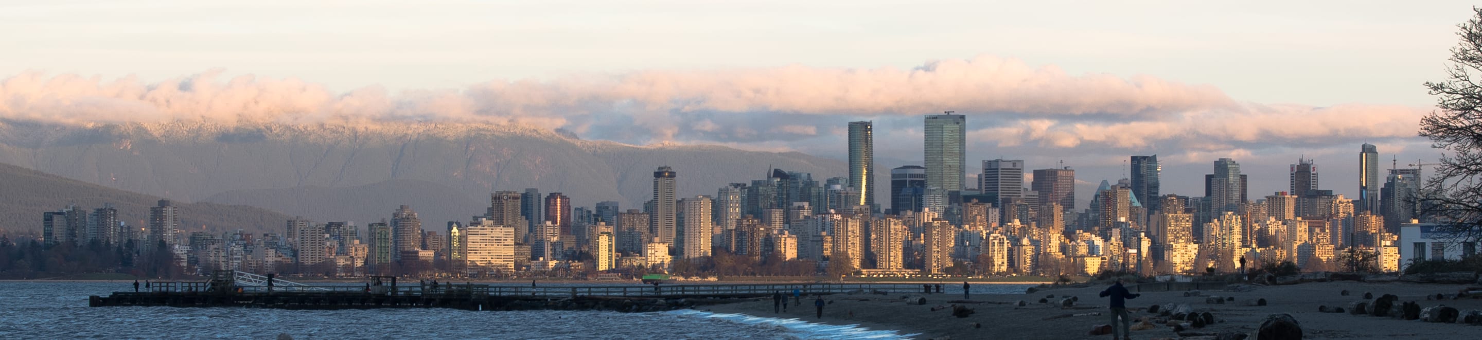 This is a panoramic photograph of a city skyline at sunset, with mountains and clouds in the background, and a beach in the foreground.