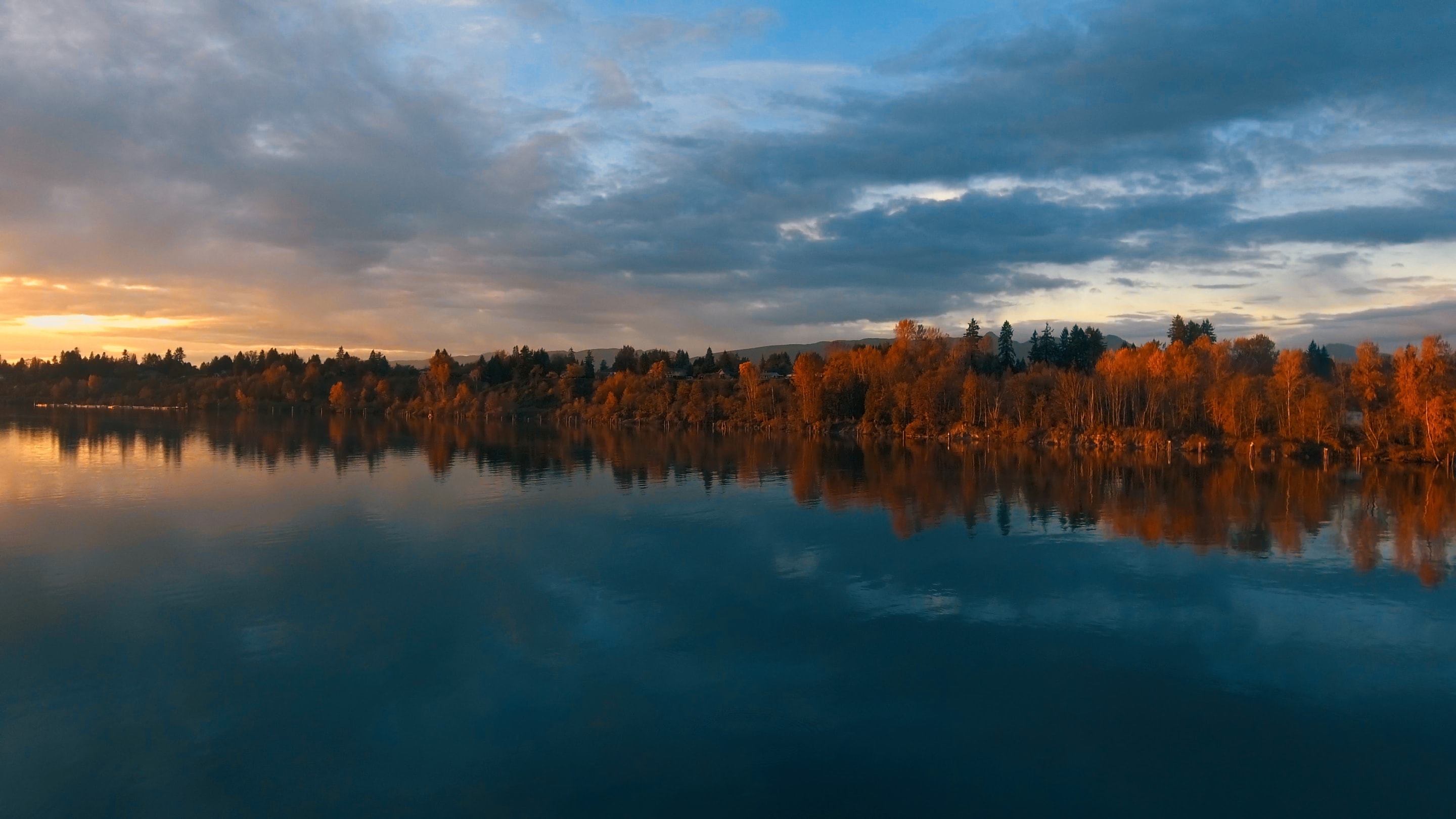 A tranquil landscape captures the vibrant colors of autumn reflected in the calm water. The sky is filled with dramatic clouds, creating a stunning contrast with the water's stillness.